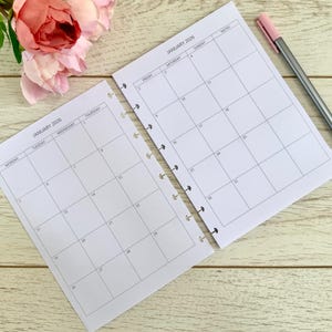 May include: Two open planner pages for January 2026 with a pink pen and faux pink and peach roses on a light wood grain surface. The planner pages are blank with a grid layout for daily scheduling.