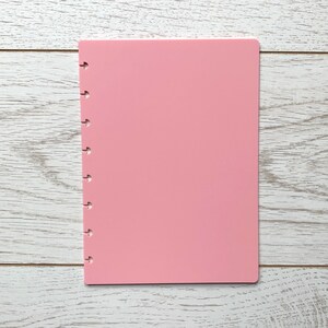 May include: A pink notebook with a hole punch on the left side. The notebook is on a white wooden surface.
