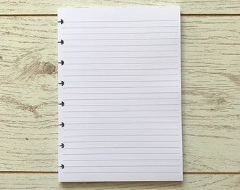 Peut inclure: Un carnet blanc à lignes avec une reliure noire et des trous sur le côté gauche. Le carnet est posé à plat sur une surface en bois clair.