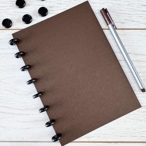 May include: A brown notebook with black binding rings and a pen with a brown cap. Several black disc-shaped objects are scattered near the notebook on a white wooden surface. The notebook is open and ready for use.