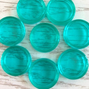 May include: Eight teal-coloured, round glass pieces are arranged in a circle. Each piece has a slightly raised edge and a textured, wood-grain appearance. They are set on a light-coloured, wood-grain surface.