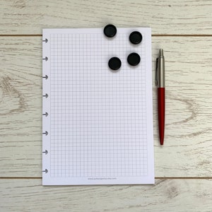 May include: A white gridded notebook with a black and white website address printed at the bottom. Four black magnetic disc-shaped page holders are placed on the top of the notebook. A red pen is placed to the right of the notebook.