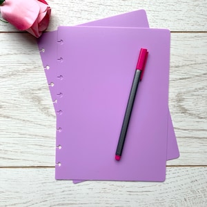 May include: Two purple notebooks with a black pen and a pink rose on a white wooden surface.