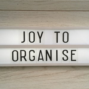 A white light-up sign with black letters that reads 'JOY TO ORGANISE' with a black heart on each side of the sign.
