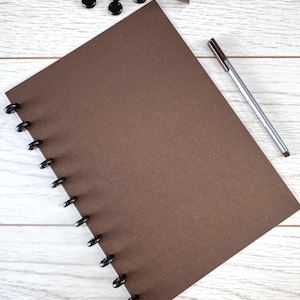 May include: A brown disc-bound notebook with black discs, a silver pen, black round magnets, and a roll of brown tape on a white wooden surface. The notebook is open, displaying a blank page. The pen has a brown ink cartridge.