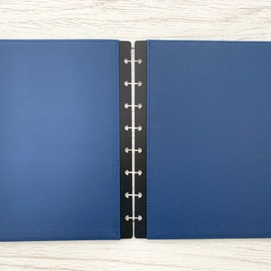 A dark blue faux leather Discbound planner cover with black plastic mushroom punched edging. The planner cover is open and lying on a white wooden surface.