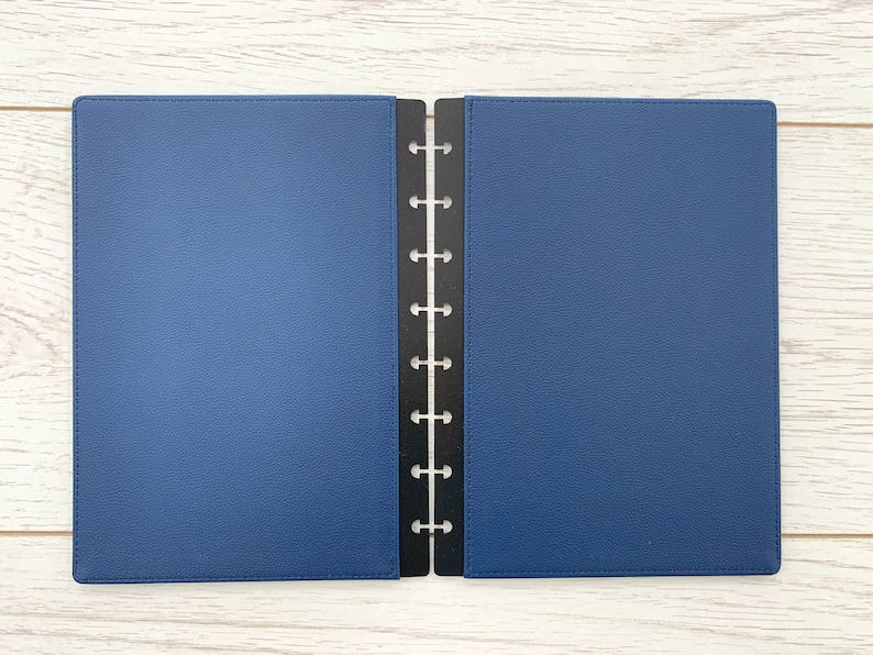 A dark blue faux leather Discbound planner cover with black plastic mushroom punched edging. The planner cover is open and lying on a white wooden surface.