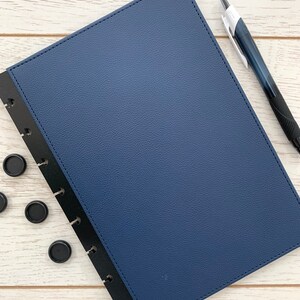 A dark blue faux leather bound notebook cover with black plastic mushroom punched edging. The notebook cover is closed and is lying on a white wooden surface with a black pen and some black binding discs lying next to it.