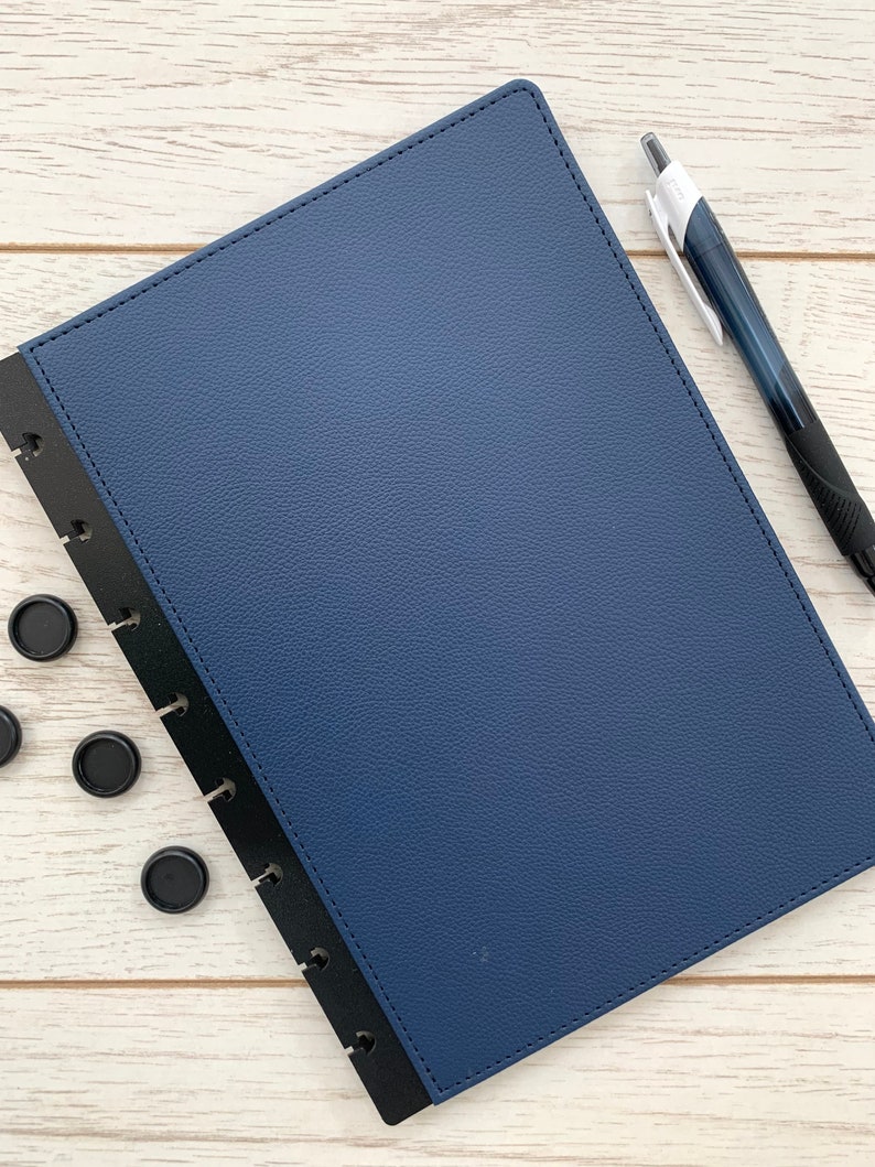 A dark blue faux leather bound notebook cover with black plastic mushroom punched edging. The notebook cover is closed and is lying on a white wooden surface with a black pen and some black binding discs lying next to it.