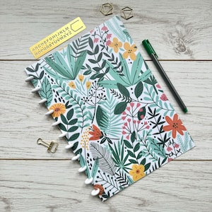 May include: A white notebook with a floral pattern featuring green leaves, yellow, orange, and pink flowers. The notebook has a white coil binding and a gold alphabet stencil on top.