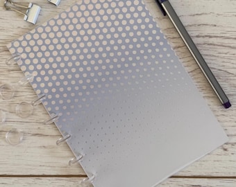 May include: A notebook with a light blue and white polka dot pattern that fades to white. A silver pen and two silver binder clips are on the wooden surface. Clear disc-bound rings are visible on the left side.