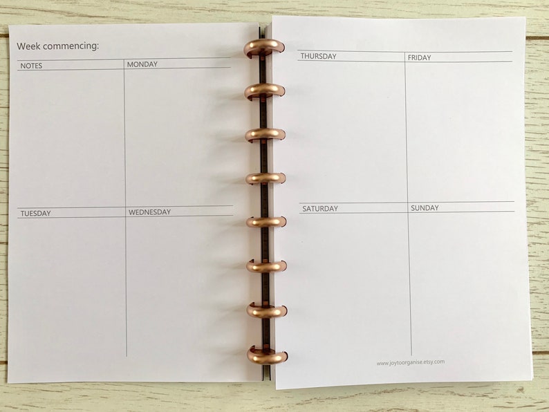 May include: A white weekly planner with a black and white grid layout. The planner has a section for notes, Monday through Sunday, and a space for the week commencing date. The planner is bound with a gold metal coil.