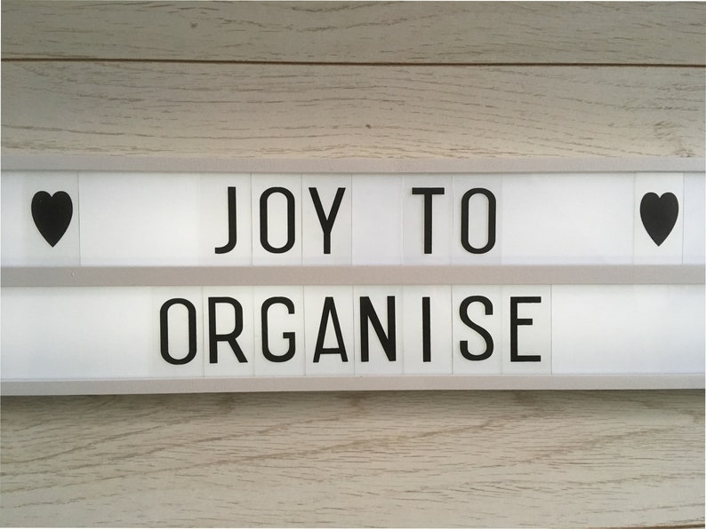 A white light-up sign with black letters that reads 'JOY TO ORGANISE' with a black heart on each side of the sign.