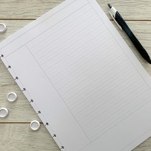 May include: A white notebook with a black pen on a white wooden surface. The notebook has a hole punch on the left side and lined pages. There are several white round objects scattered around the notebook.