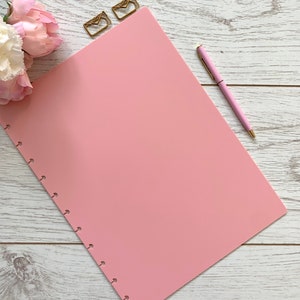 May include: A pink planner with a hole punch design and a pink pen on a white wooden surface. There are two gold paper clips in the shape of envelopes on the top of the planner.