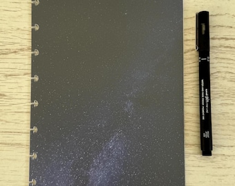 May include: A dark grey planner insert with a starry design, featuring holes along the left edge for binding. A black fine-line pen is positioned to the right, with the text "Uni Pin Fine Line" visible.