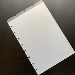 May include: A translucent white planner divider with four tabs. The divider has a series of holes along the left edge for binding. The tabs are slightly darker than the main body of the divider. The divider is on a dark surface.