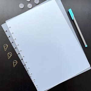 May include: A stack of light blue, translucent binder pages with a row of holes along the left edge. A silver pen with a teal tip and a few gold paper clips are also present. Small, clear plastic discs are scattered at the top.
