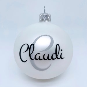 Könnte beinhalten: Weißer Weihnachtsschmuck mit dem Namen "Claudi" in schwarzer Schrift, mit einem silbernen Buchstaben "C" darüber. Der Schmuck hat eine silberne Metallkappe und einen Haken zum Aufhängen.
