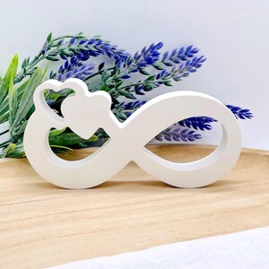 Könnte beinhalten: Ein weißes Holz-Unendlichkeitszeichen mit einem Herz-Design. Das Symbol steht auf einer Holzoberfläche, im Hintergrund lila und grünes Laub. Das Unendlichkeitszeichen ist ein dekorativer Artikel.