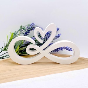Könnte beinhalten: Ein weißes Holz-Unendlichkeitszeichen mit Herzdesign. Das Symbol befindet sich auf einem Holztablett mit Zweigen lila Blumen im Hintergrund. Das Symbol ist ein dekorativer Gegenstand.