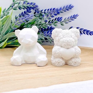 Könnte beinhalten: Zwei weiße Teddybär-Figuren. Einer ist ein geometrisches Design, der andere ist mit rosenartigen Details bedeckt und hält ein Herz. Die Bären stehen auf einem Holztablett mit lila und grünen floralen Akzenten.