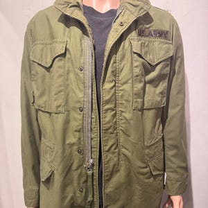 May include: Olive green military-style jacket with a full zipper closure and multiple front pockets. The jacket features a collar and the text "US ARMY" on the upper right chest. The jacket is displayed on a mannequin.