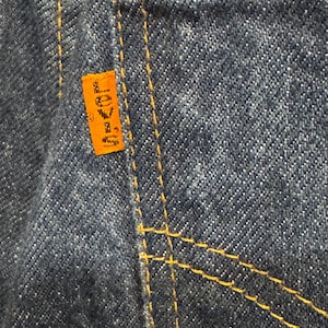 May include: A close-up of blue denim fabric featuring thick, golden-yellow contrast stitching along the seams. An orange rectangular fabric tab is sewn into the side seam, displaying the word Levi's in black lettering with a small registered trademark symbol.