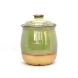 May include: A handmade ceramic jar with a lid. The jar is primarily light green with a band of tan at the base. The lid is a darker shade of green. The jar has a glossy finish and is likely used for storage.