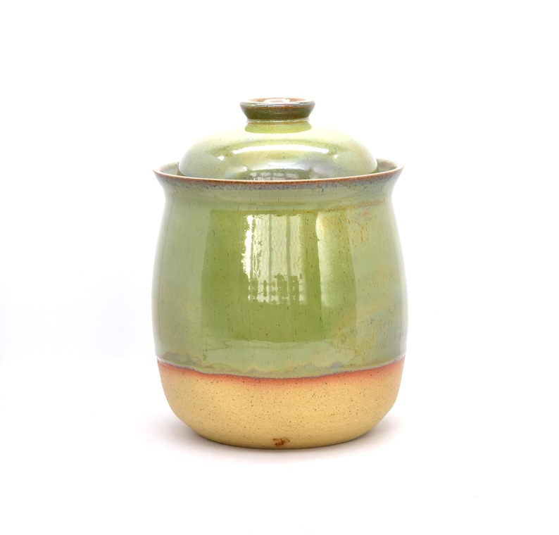 May include: A handmade ceramic jar with a lid. The jar is primarily light green with a band of tan at the base. The lid is a darker shade of green. The jar has a glossy finish and is likely used for storage.