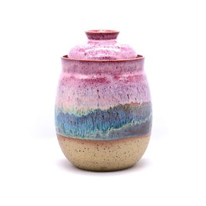 May include: A handmade ceramic jar with a lid. The jar features a speckled beige base, transitioning to bands of blue, purple, and pink hues. The lid is a matching pink color. The jar is a decorative storage container.