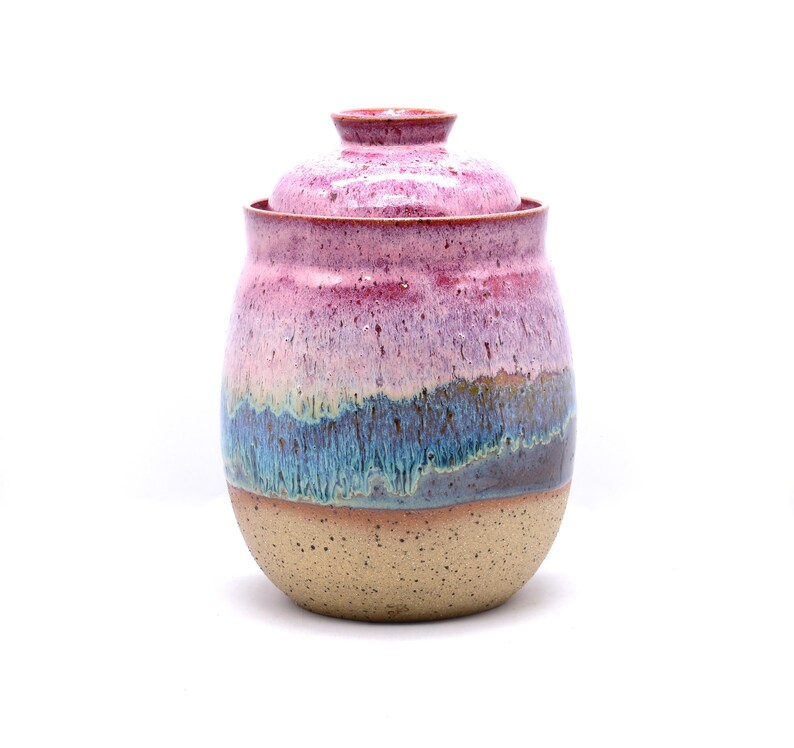 May include: A handmade ceramic jar with a lid. The jar features a speckled beige base, transitioning to bands of blue, purple, and pink hues. The lid is a matching pink color. The jar is a decorative storage container.