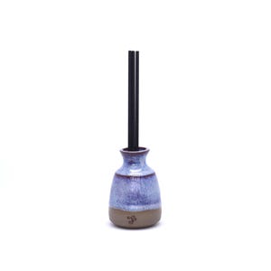 May include: A ceramic reed diffuser vessel with a rounded base and a narrow neck. The upper portion features a mottled light blue and purple glaze, while the lower base is unglazed brown clay with a small stamped letter F. Three black reeds.