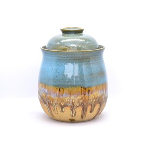 May include: A ceramic jar with a lid, featuring a blue and tan color scheme. The jar has a rounded shape with a lid on top. The upper portion is blue, transitioning to tan with a decorative pattern near the base. The jar is likely handmade.