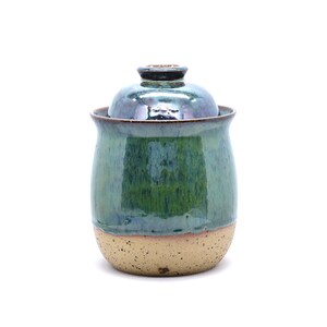 May include: A handmade ceramic jar with a lid. The jar is glazed in shades of green and blue, with a speckled tan base. The lid is a darker shade of blue-green. The jar has a rounded shape and a glossy finish.