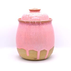 May include: A handmade ceramic jar with a lid. The jar is a soft pink color with a dripped glaze effect over a tan base. The lid is also pink and fits snugly on top. The jar is a decorative piece.