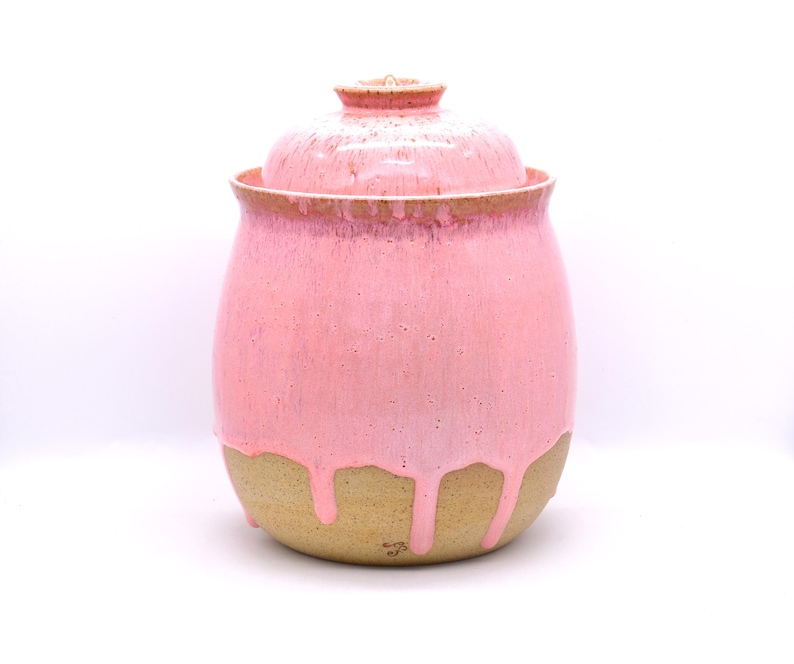 May include: A handmade ceramic jar with a lid. The jar is a soft pink color with a dripped glaze effect over a tan base. The lid is also pink and fits snugly on top. The jar is a decorative piece.