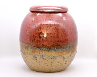 May include: A ceramic jar with a lid, featuring a glossy, reddish-brown upper half and a speckled, tan lower half. The jar has a rounded shape with a slight taper towards the base. The glaze has a dripped effect where the colors meet.