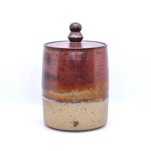 Puede incluir: Un tarro de cerámica hecho a mano con tapa. El tarro tiene forma cilíndrica con una base beige moteada y una sección superior marrón rojizo. La tapa es de color marrón rojizo a juego con un pequeño pomo en la parte superior. El tarro probablemente se utiliza para el almacenamiento.