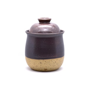 May include: A handmade ceramic jar with a lid. The jar is a deep plum color with a speckled tan base. The lid has a small knob on top. The jar is likely used for storing food items.