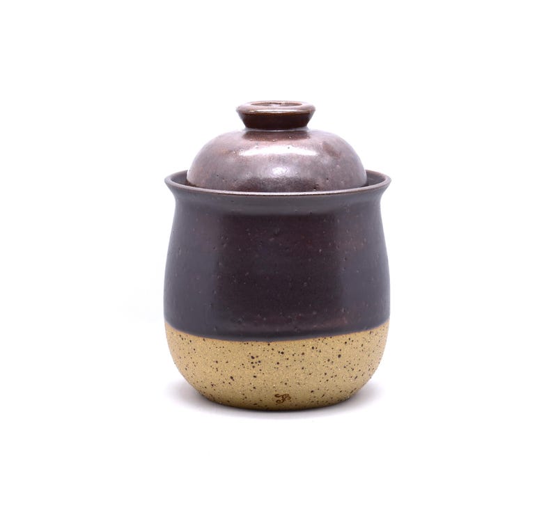 May include: A handmade ceramic jar with a lid. The jar is a deep plum color with a speckled tan base. The lid has a small knob on top. The jar is likely used for storing food items.