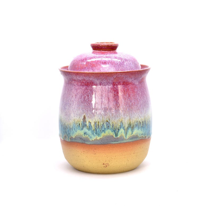 May include: A handmade ceramic jar with a lid. The jar has a rounded shape and is glazed in a gradient of colors, including pink, purple, blue, and yellow. The lid is a matching pink color. The jar is likely used for storage.