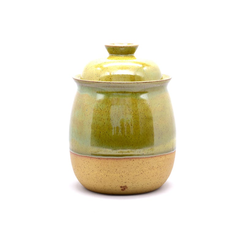 May include: A handmade ceramic jar with a lid. The jar is a light green color with a speckled tan base. The lid has a small knob on top. The jar is likely used for storage.