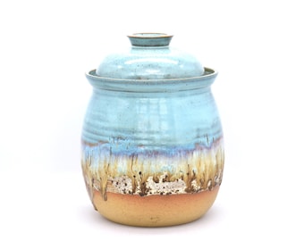 May include: A ceramic canister with a lid, featuring a light blue glazed upper section that transitions into a drippy, multi-colored glaze of white, brown, and tan, ending in a solid, unglazed matte tan base with a rounded, bulbous shape.