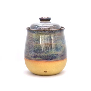 May include: A handmade ceramic jar with a lid. The jar has a rounded shape with a yellow base, transitioning to blue and green hues towards the top. The lid is a darker shade of blue. A small signature is visible on the base.
