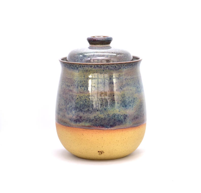 May include: A handmade ceramic jar with a lid. The jar has a rounded shape with a yellow base, transitioning to blue and green hues towards the top. The lid is a darker shade of blue. A small signature is visible on the base.