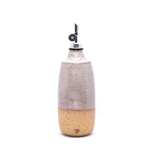 May include: A ceramic oil dispenser with a two-tone speckled finish. The top half is a light mauve-grey, while the bottom half is a warm tan. It features a silver-colored metal pour spout with a black rubber stopper and a small dark logo mark.