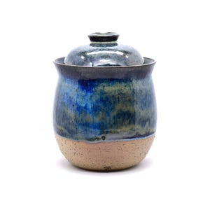 May include: A ceramic jar with a lid featuring a glossy glaze in shades of deep blue, navy, and muted green. The lower portion of the jar is made of unglazed, speckled tan clay with a rounded base and a slightly flared rim at the top opening.