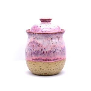 May include: A ceramic jar with a lid, featuring a speckled tan base and a body with a gradient of pink and purple hues. The jar has a rounded shape and a matching lid with a small knob.