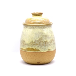 May include: A handmade ceramic jar with a lid. The jar has a rounded shape with a light brown base and a creamy, off-white upper section. The lid is a matching color. The jar is suitable for kitchen storage.